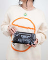 Person wearing a cream sweater with a large orange circle design, holding a black wallet with brand logo.