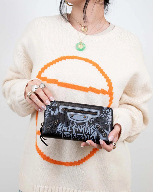 Person wearing a cream sweater with a large orange circle design, holding a black wallet with brand logo.