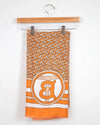 Orange and beige patterned scarf with a logo on a hanger against a white background