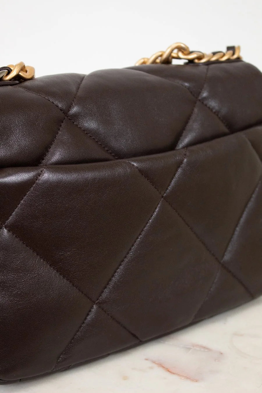 Close-up of a dark brown quilted handbag with gold chain handle on a light marble surface.