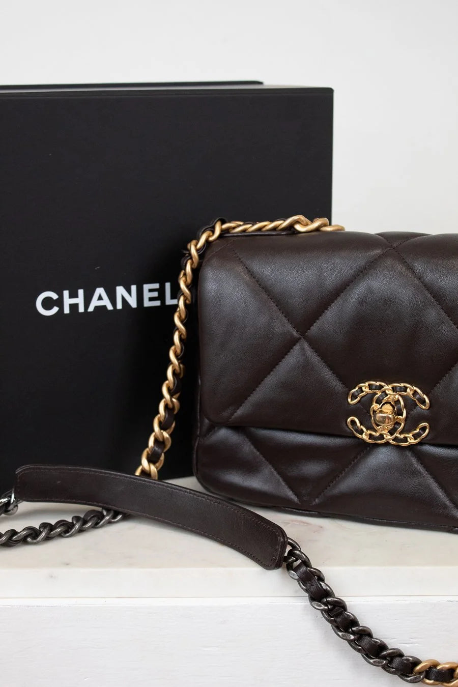 Dark Brown quilted Chanel handbag with gold chain and clasp on a white surface, with a black Chanel box in the background.