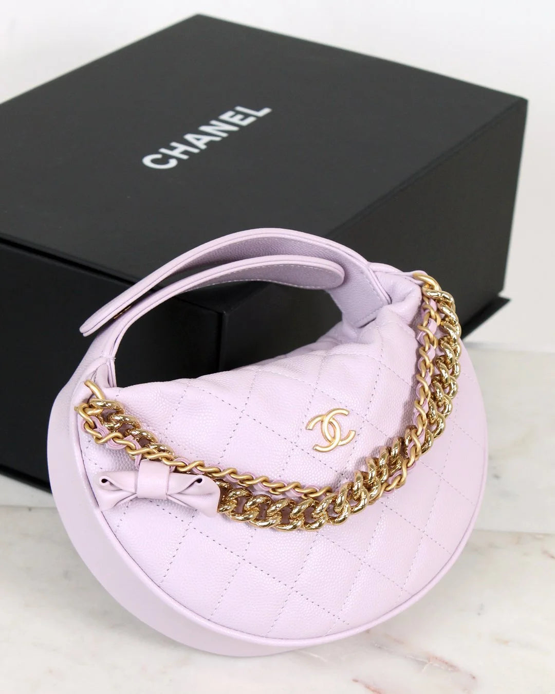Lilac bag with gold chain and Chanel logo in front of a black Chanel box.