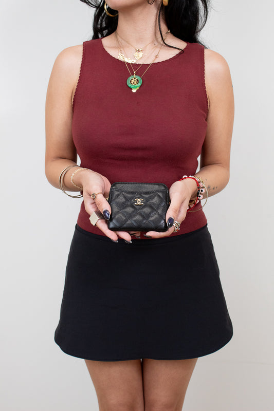 Person wearing a burgundy top and black skirt holding a black Chanel wallet against a plain background