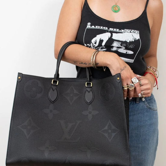 Person holding a black handbag with a visible brand logo, wearing a black tank top and blue jeans.