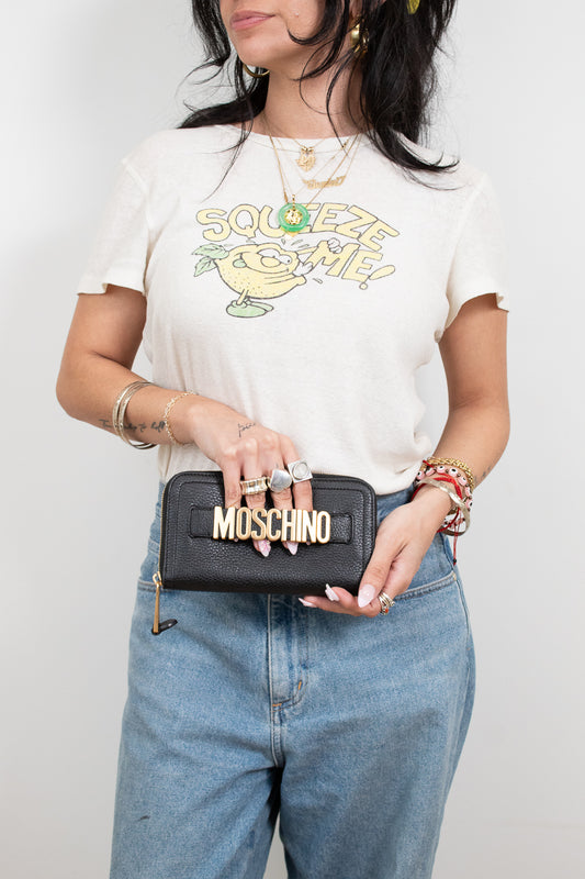 Person wearing a white t-shirt with a graphic design, holding a black wallet with 'Moschino' branding.