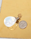 Gold pendant with a large white stone next to a silver coin on a gold plate background for size reference