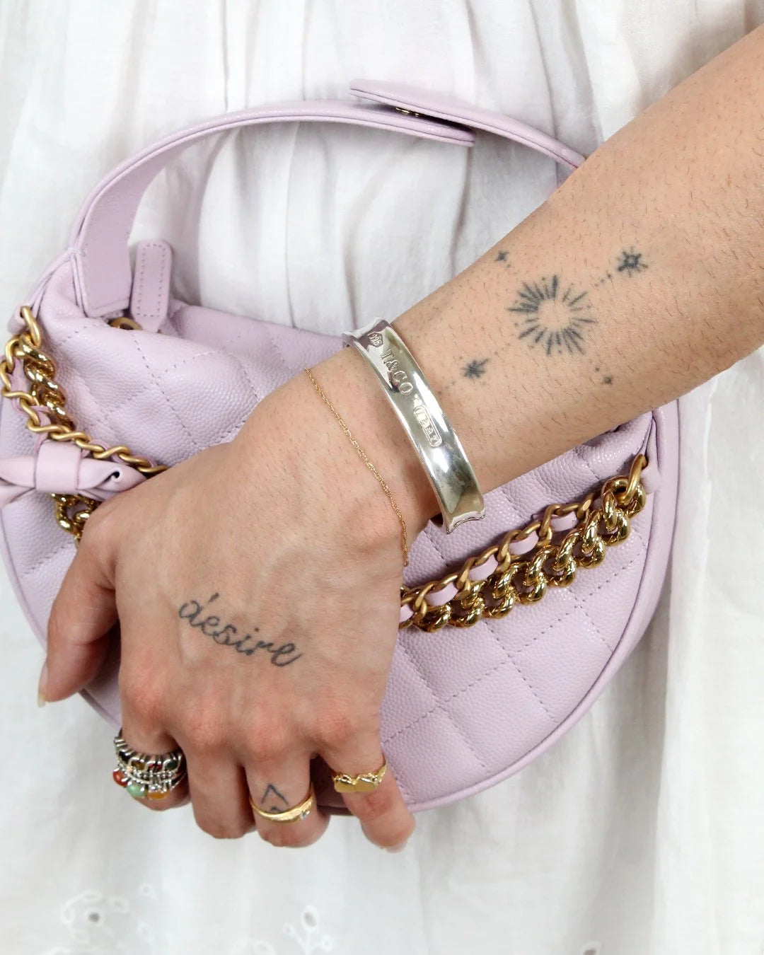 Hand with tattoos holding a pink quilted handbag with gold chain strap and wearing a silver cuff