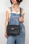 Person wearing denim overalls holding a black studded handbag against a plain background