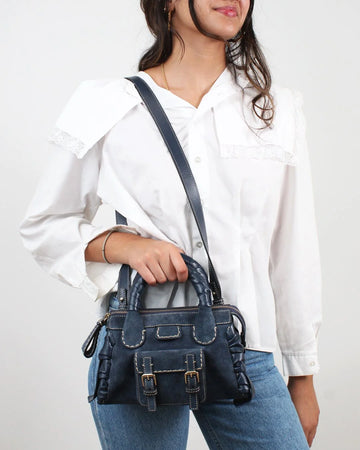 Person wearing a white shirt and blue jeans with a dark blue handbag on a plain background