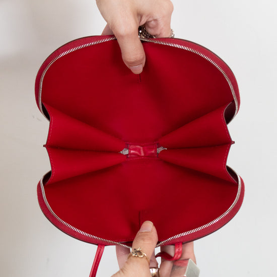 A red handbag  with a unique heart-like layered design and opened silver zipper, held open by two hands against a plain white background.