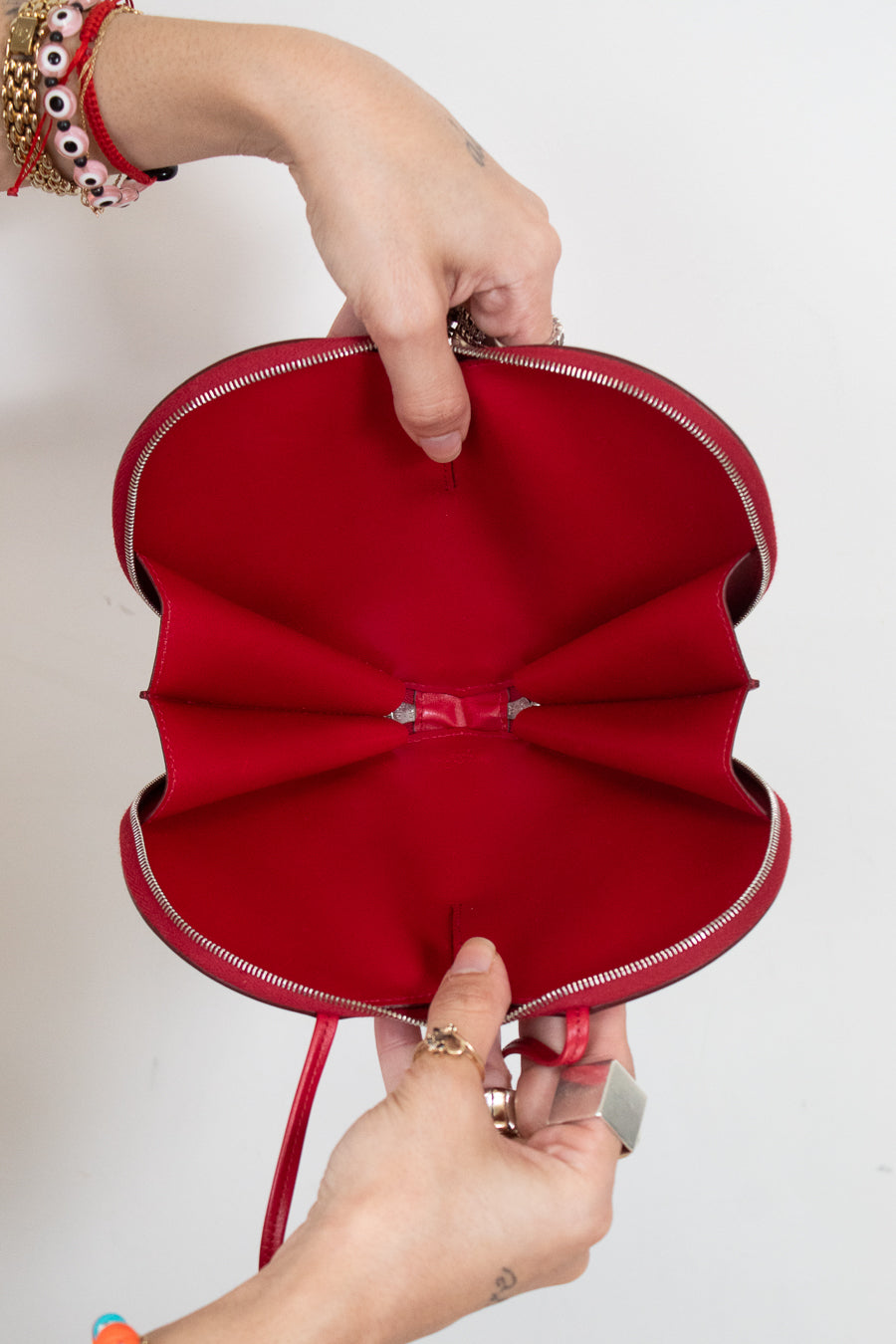 A red handbag  with a unique heart-like layered design and opened silver zipper, held open by two hands against a plain white background.