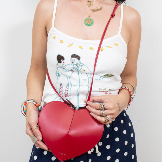 A woman wearing a white spaghetti-strap tank top with a cartoon couple illustration and navy blue polka-dot pants, holding a red heart-shaped leather crossbody bag with a red strap.