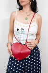 A woman wearing a white spaghetti-strap tank top with a cartoon couple illustration and navy blue polka-dot pants, holding a red heart-shaped leather crossbody bag with a red strap.