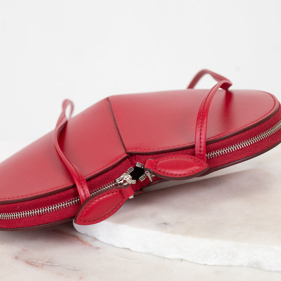 A red leather zippered handbag with thin straps and a silver zipper, resting on a white marble slab against a white background.