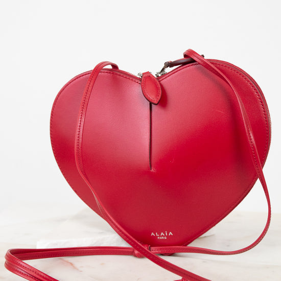 A bright red heart-shaped leather crossbody bag with a matching strap, displayed on a light marble surface against a white background. The brand name 'ALAIA PARIS' is printed in gold near the bottom of the bag.