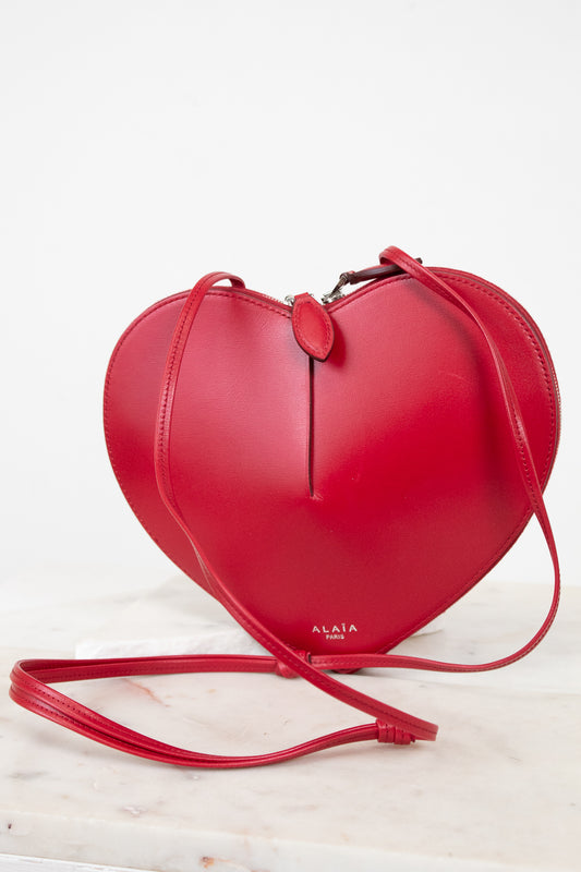 A bright red heart-shaped leather crossbody bag with a matching strap, displayed on a light marble surface against a white background. The brand name 'ALAIA PARIS' is printed in gold near the bottom of the bag.