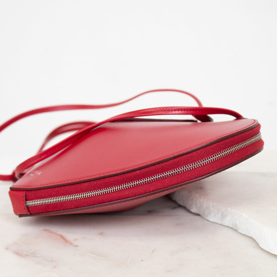 A red leather crossbody bag with a thin strap and silver zipper, resting on a white marble surface against a white background.