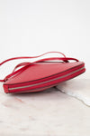 A red leather crossbody bag with a thin strap and silver zipper, resting on a white marble surface against a white background.