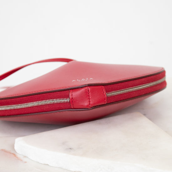 A red leather clutch bag with a silver zipper and a small embossed brand name, resting on a white marble slab against a white background.