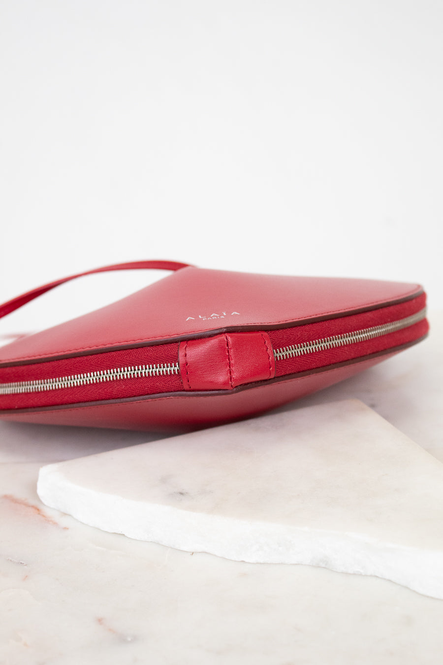 A red leather clutch bag with a silver zipper and a small embossed brand name, resting on a white marble slab against a white background.