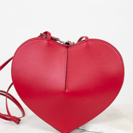 A bright red heart-shaped leather crossbody bag with a matching red strap, resting on a white marble surface against a white background.