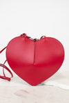 A bright red heart-shaped leather crossbody bag with a matching red strap, resting on a white marble surface against a white background.