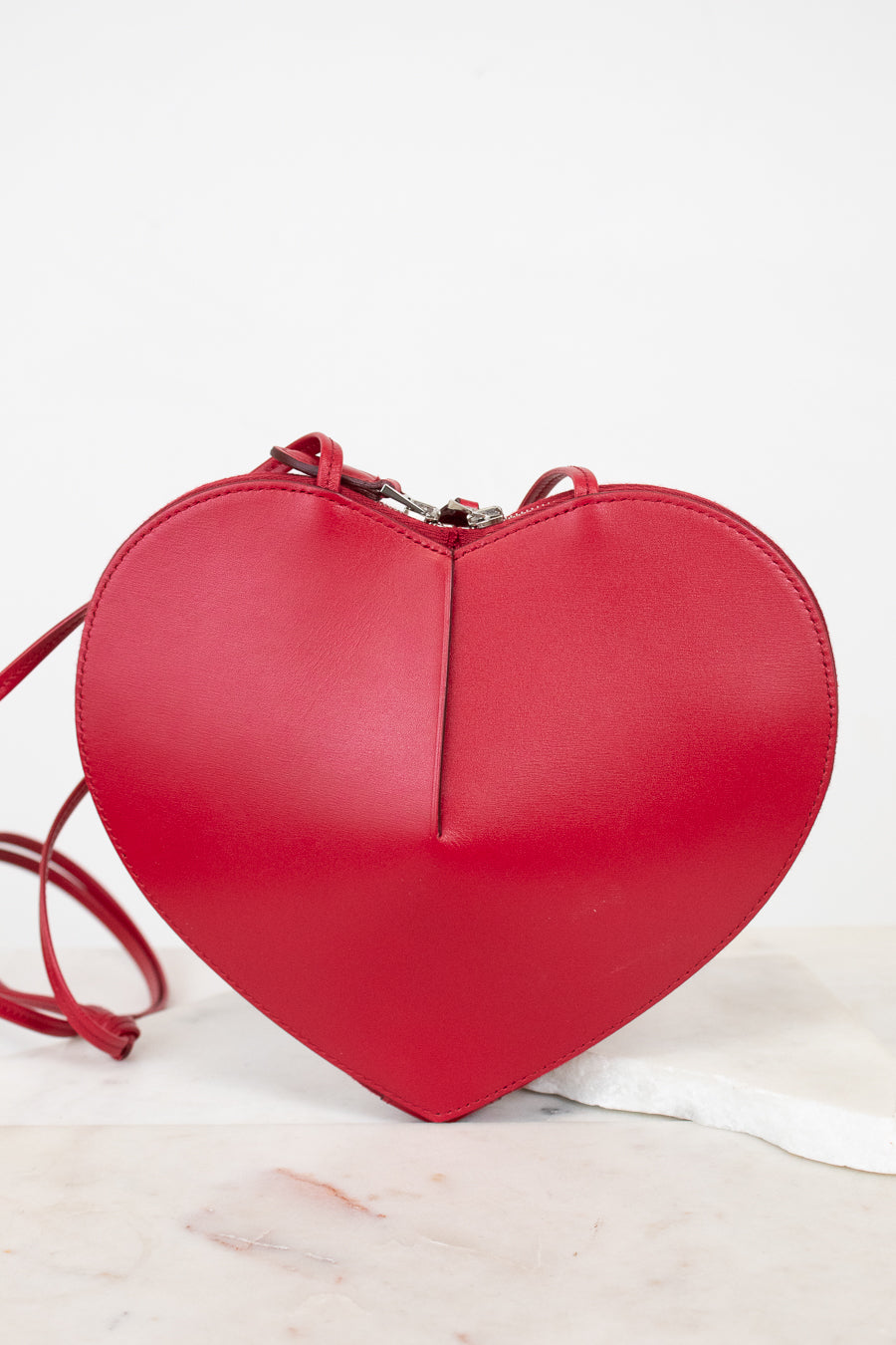 A bright red heart-shaped leather crossbody bag with a matching red strap, resting on a white marble surface against a white background.