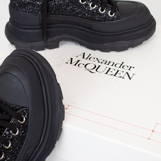 A pair of black chunky lace-up boots with glittery uppers and thick black soles, displayed on a white box printed with the brand name Alexander McQueen.
