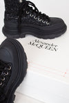 A pair of black chunky lace-up boots with glittery uppers and thick black soles, displayed on a white box printed with the brand name Alexander McQueen.