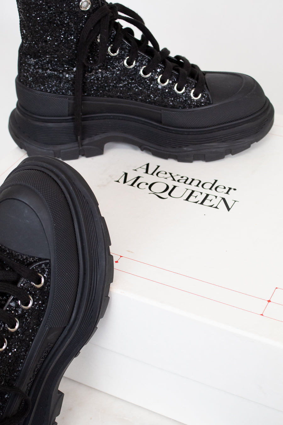 A pair of black chunky lace-up boots with glittery uppers and thick black soles, displayed on a white box printed with the brand name Alexander McQueen.