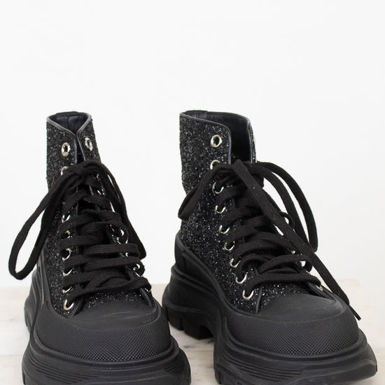 A pair of black lace-up combat boots with a glittery textured upper and chunky black soles, displayed on a white marble surface against a white background.