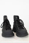 A pair of black lace-up combat boots with a glittery textured upper and chunky black soles, displayed on a white marble surface against a white background.