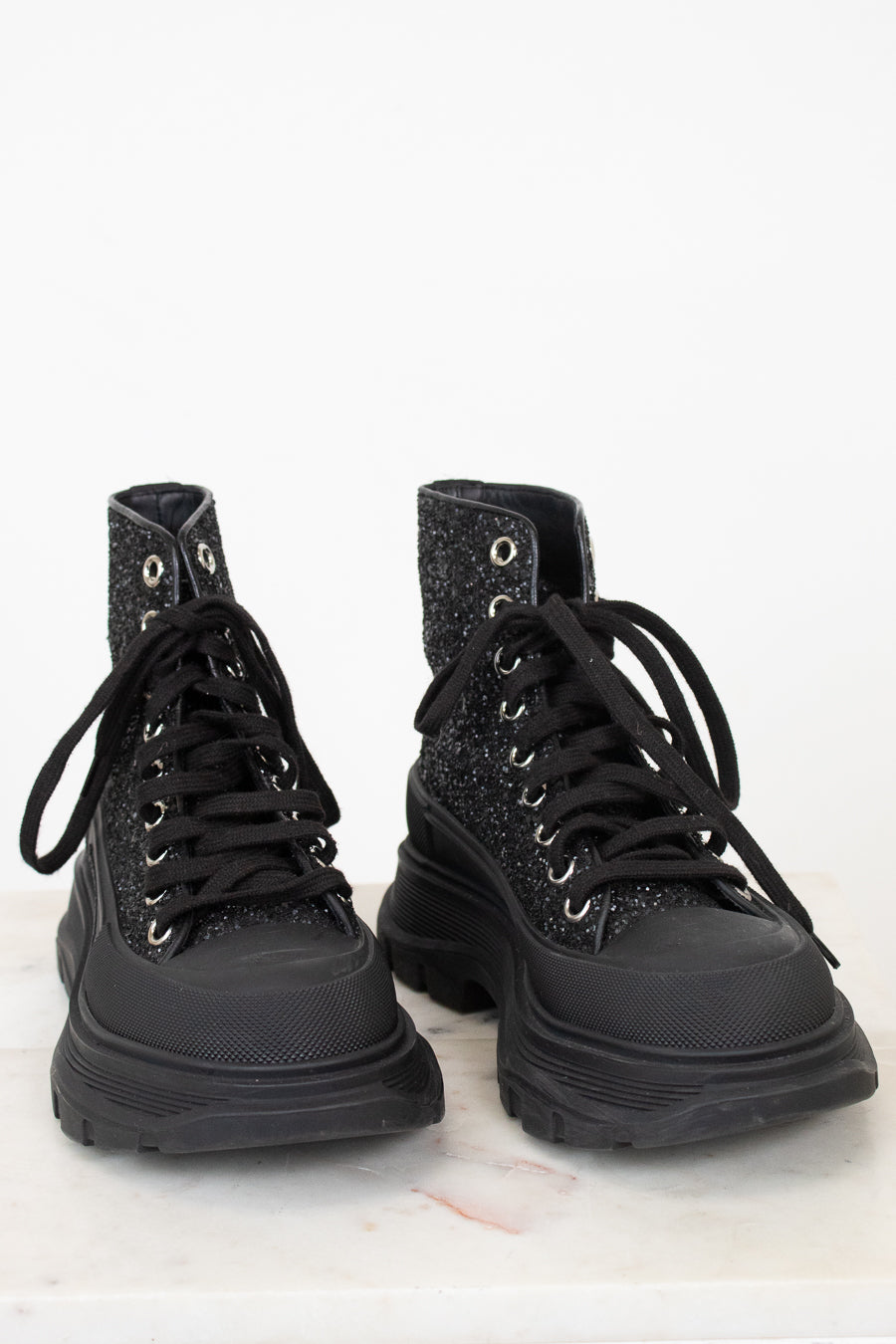 A pair of black lace-up combat boots with a glittery textured upper and chunky black soles, displayed on a white marble surface against a white background.