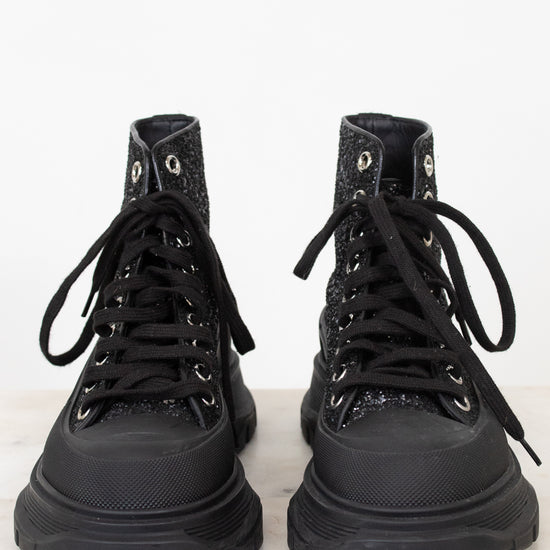 A pair of black high-top lace-up boots with textured, glittery uppers and chunky black soles, photographed from the front on a light marble surface against a white background.