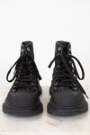 A pair of black high-top lace-up boots with textured, glittery uppers and chunky black soles, photographed from the front on a light marble surface against a white background.