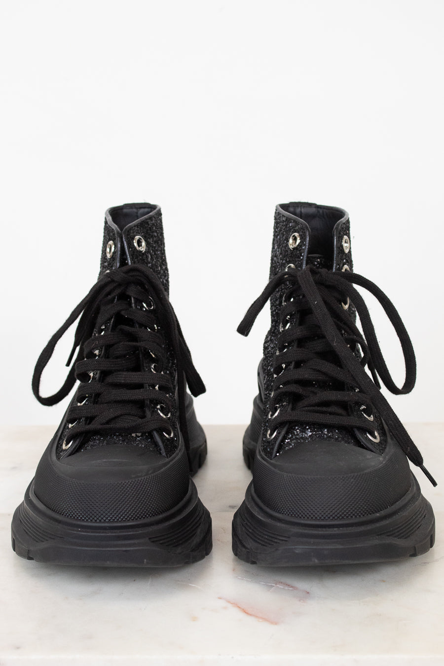 A pair of black high-top lace-up boots with textured, glittery uppers and chunky black soles, photographed from the front on a light marble surface against a white background.