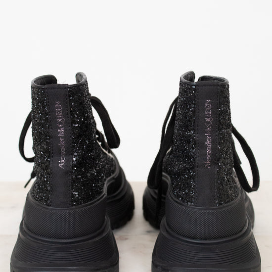 A pair of black chunky lace-up boots with glittery textured uppers, shown from the back on a light marble surface against a white background.