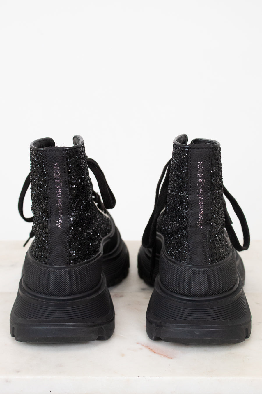 A pair of black chunky lace-up boots with glittery textured uppers, shown from the back on a light marble surface against a white background.
