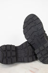 Close-up of the treaded black rubber soles of a pair of boots, showing the lug pattern and embossed branding, set against a clean white background.