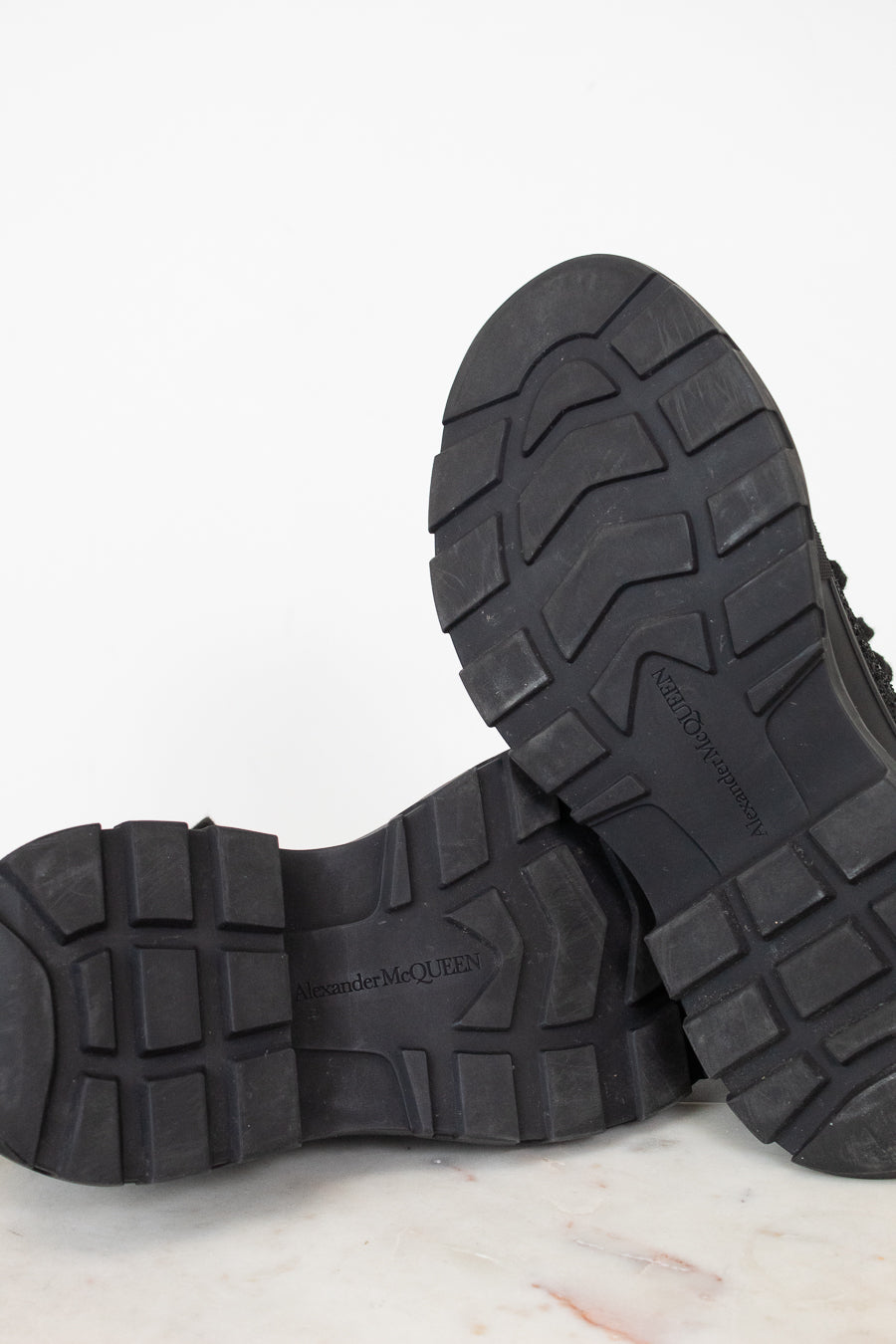 Close-up of the treaded black rubber soles of a pair of boots, showing the lug pattern and embossed branding, set against a clean white background.