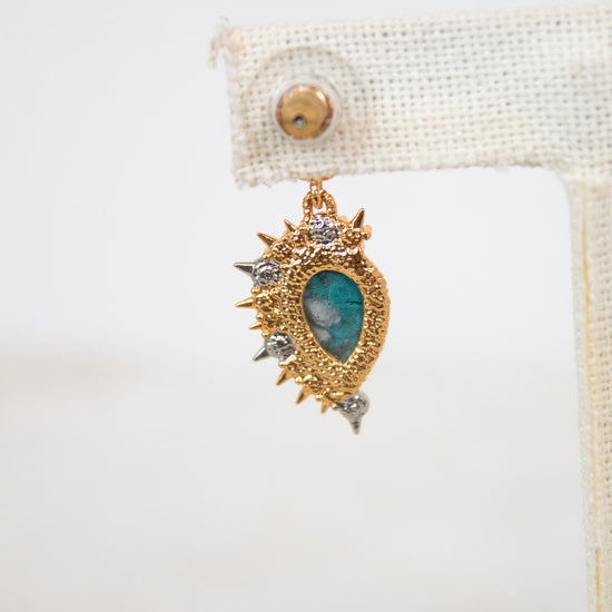 Back of gold and teal earring on a white background with a beige holder.
