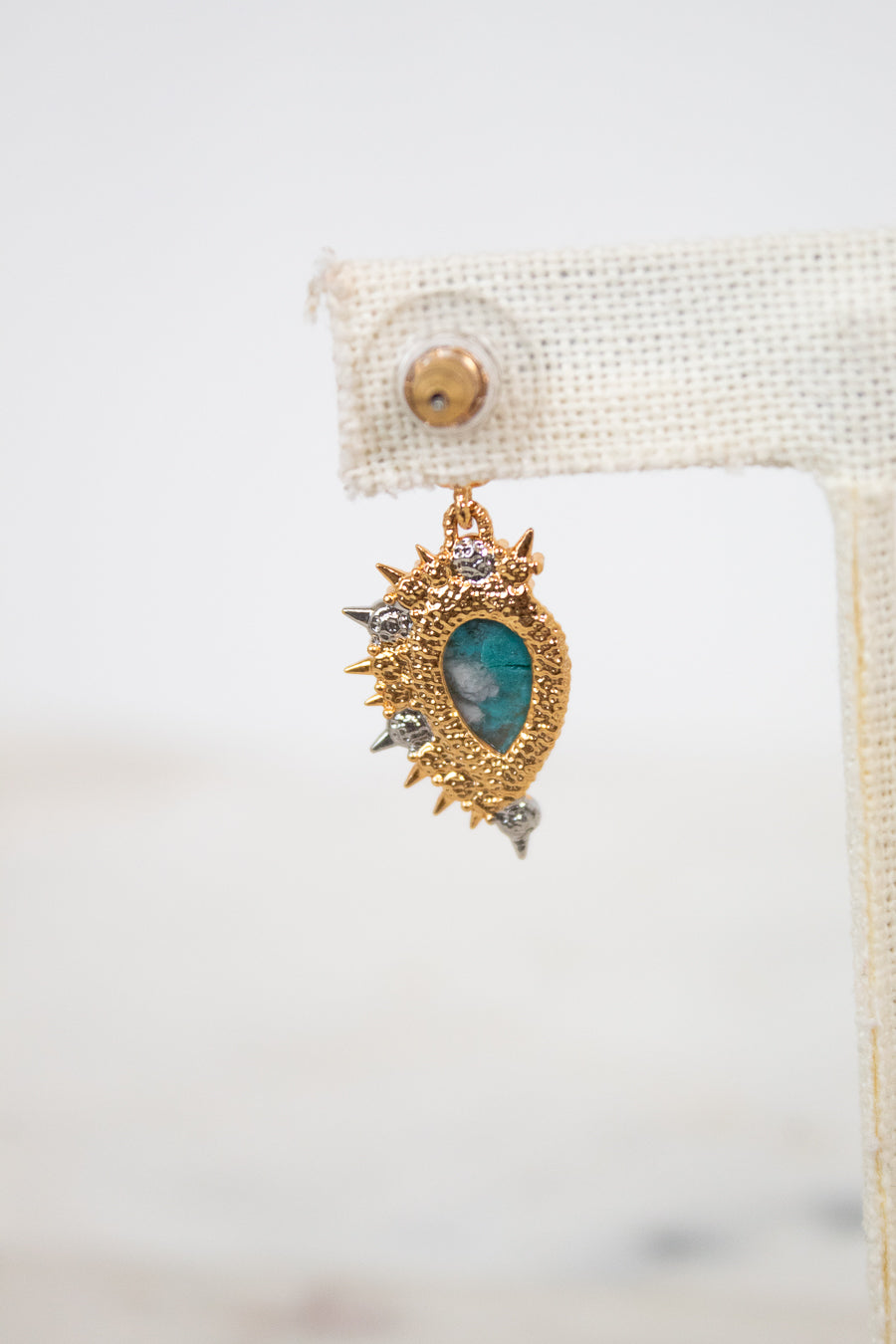 Back of gold and teal earring on a white background with a beige holder.