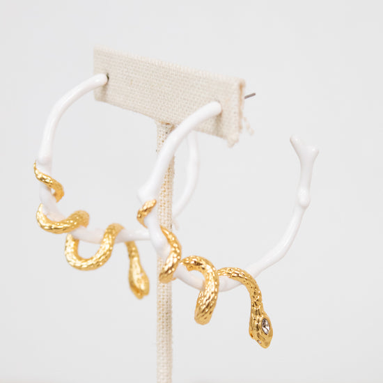 White earrings with gold snake details on a white stand against a light background