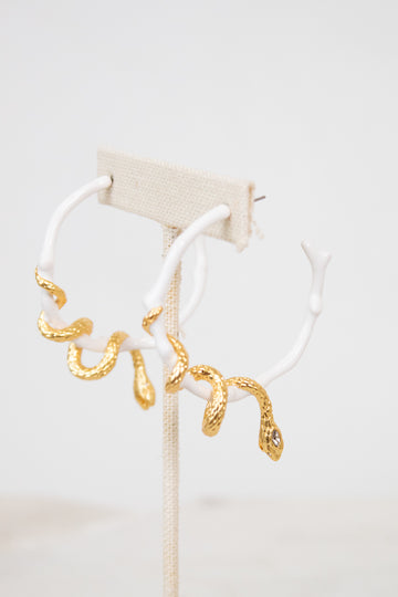 White earrings with gold snake details on a white stand against a light background
