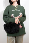 Person wearing a green sweater with a graphic design and holding a black handbag against a white background