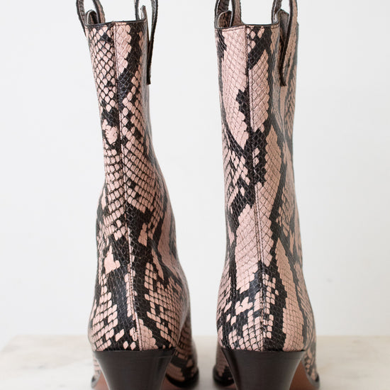 A pair of women's mid-calf heeled boots with a pink and black snakeskin pattern, shown from the back on a light marble surface against a white background.