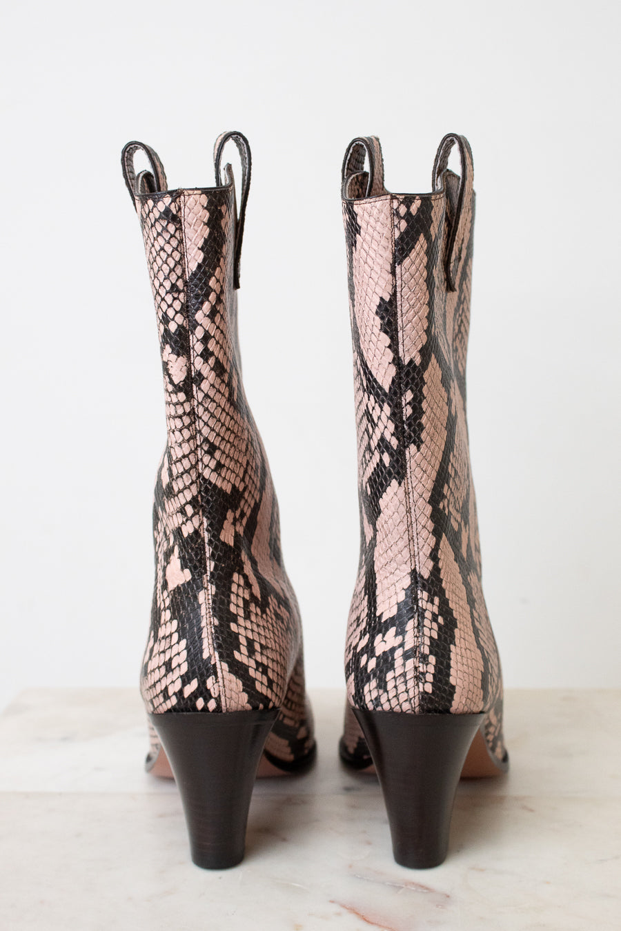 A pair of women's mid-calf heeled boots with a pink and black snakeskin pattern, shown from the back on a light marble surface against a white background.