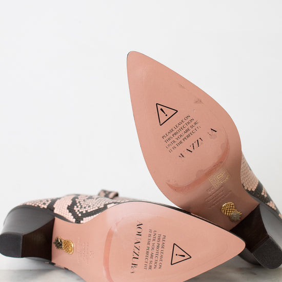 A pair of pointed-toe heeled boots with a snakeskin pattern, shown from the sole side on a light marble surface against a white background. The insole is a dusty pink color with branding and safety symbols, and the heel is dark brown with a gold accent.