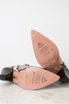A pair of pointed-toe heeled boots with a snakeskin pattern, shown from the sole side on a light marble surface against a white background. The insole is a dusty pink color with branding and safety symbols, and the heel is dark brown with a gold accent.