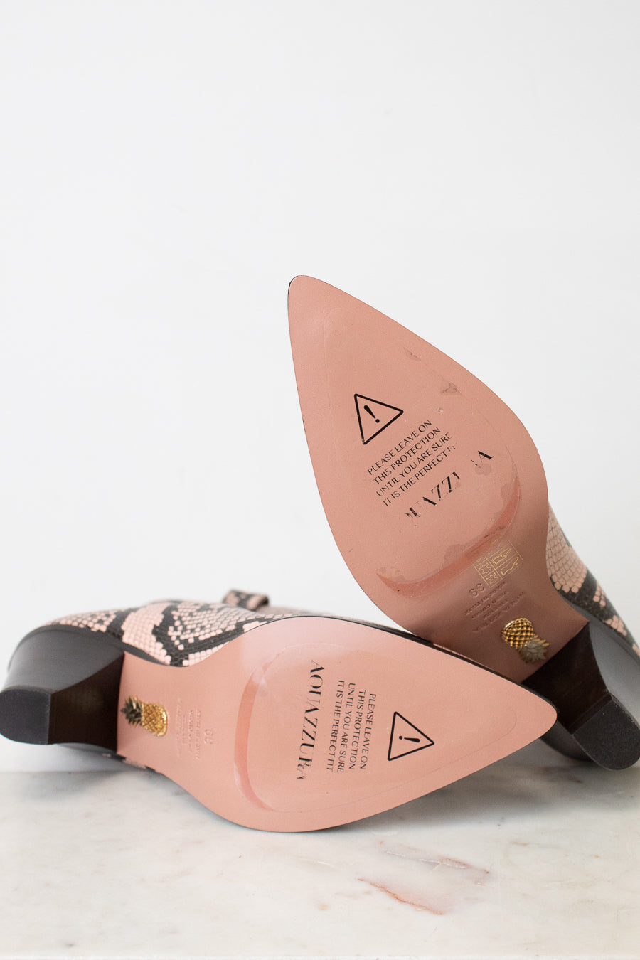 A pair of pointed-toe heeled boots with a snakeskin pattern, shown from the sole side on a light marble surface against a white background. The insole is a dusty pink color with branding and safety symbols, and the heel is dark brown with a gold accent.